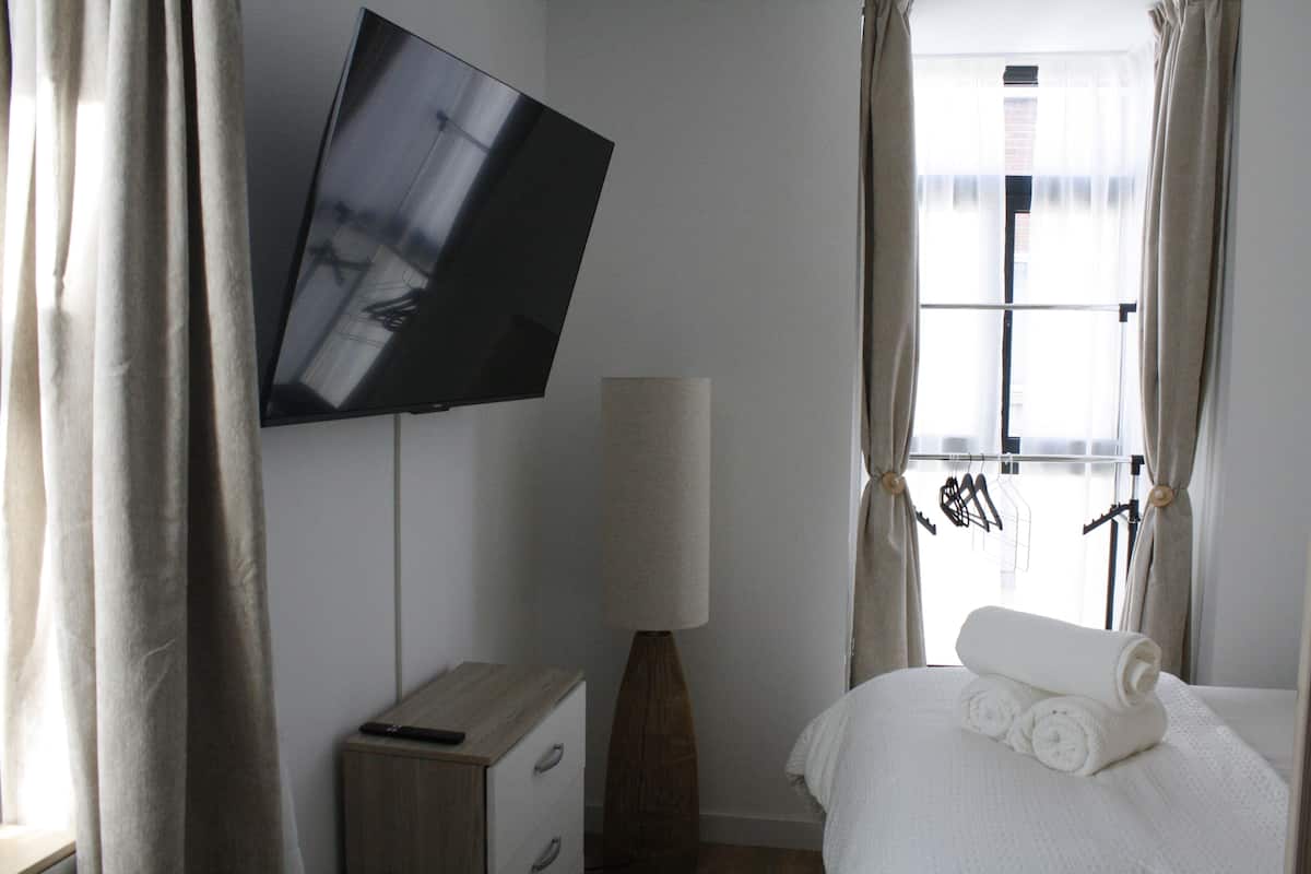 A hotel room with a flat-screen TV mounted on the wall, a bedside lamp, a neatly made bed with rolled towels, and a window with curtains.