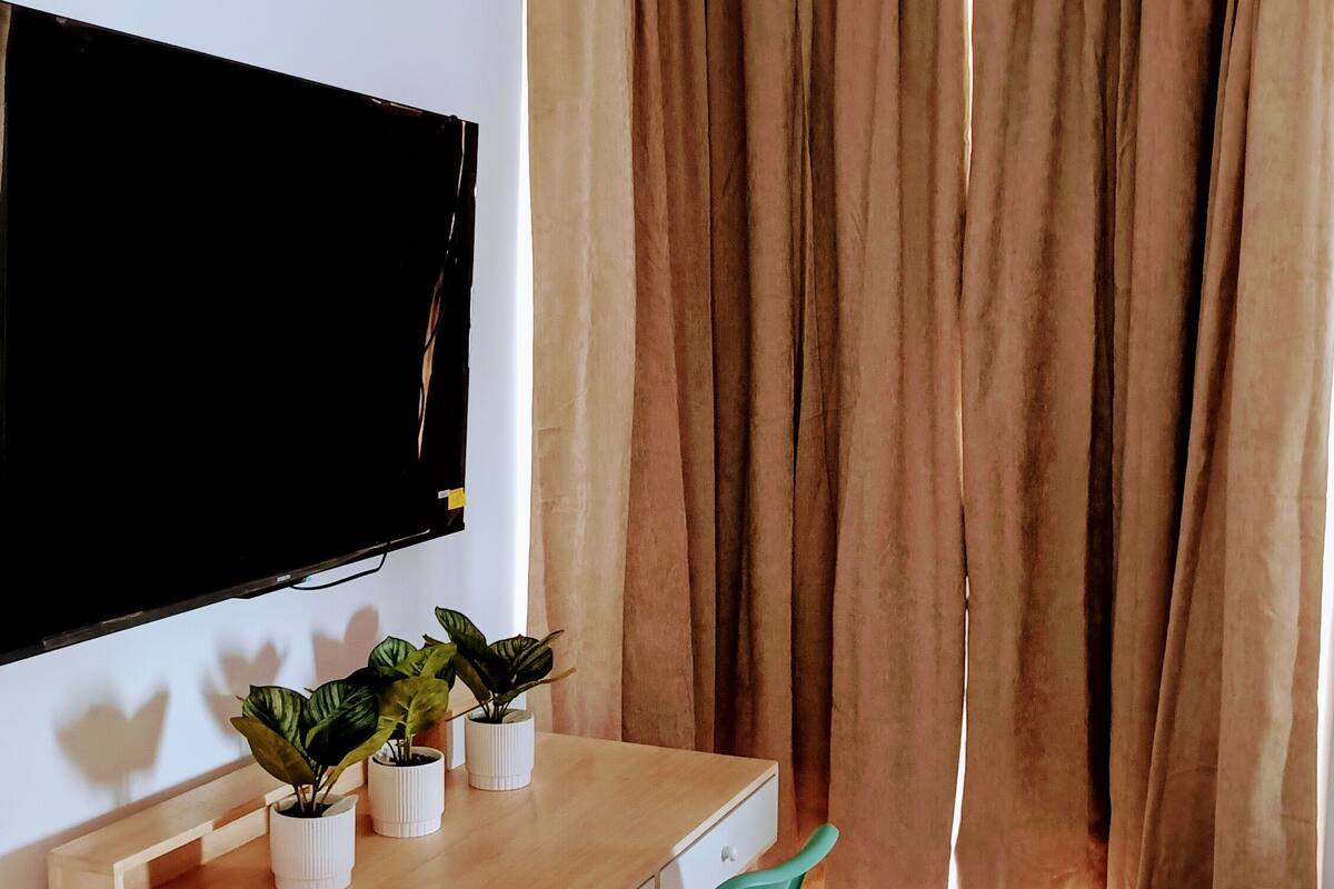 Uma sala de estar moderna com uma escrivaninha de madeira, uma poltrona verde, uma TV de tela plana na parede e uma janela grande com cortinas.