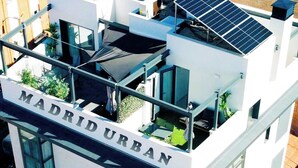 Il s’agit d’un immeuble sur lequel on peut lire l’inscription « Madrid Urban ». Il est doté d’un toit-terrasse équipé de panneaux solaires, d’un balcon agrémenté de plantes et de nombreuses fenêtres.
