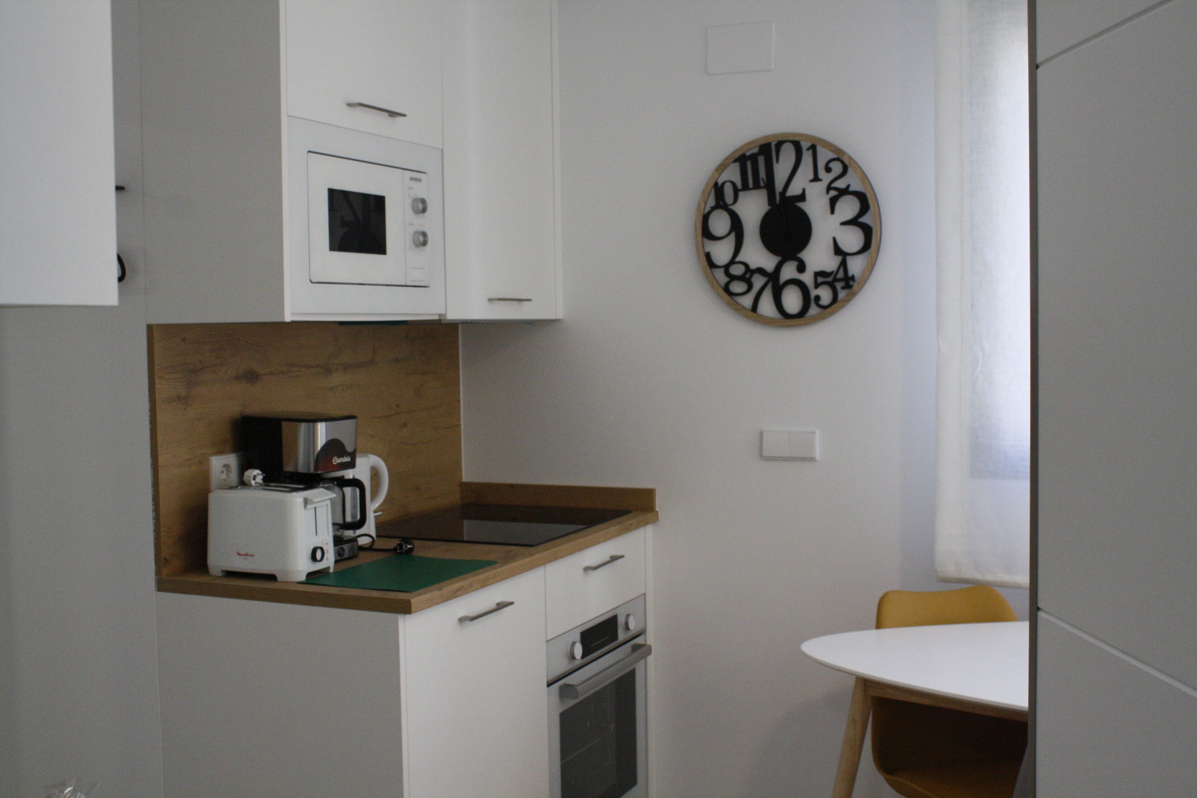 Una cocina compacta con armarios blancos, encimera de madera, cafetera y un reloj de pared.