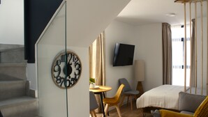 Une chambre d’hôtel moderne avec un escalier, une horloge murale ronde, un téléviseur à écran plat, un lit, un canapé et une table à manger avec des chaises.