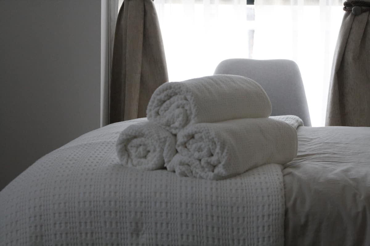 Cama bem arrumada com toalhas brancas dobradas e um travesseiro branco, cortinas e uma janela ao fundo.