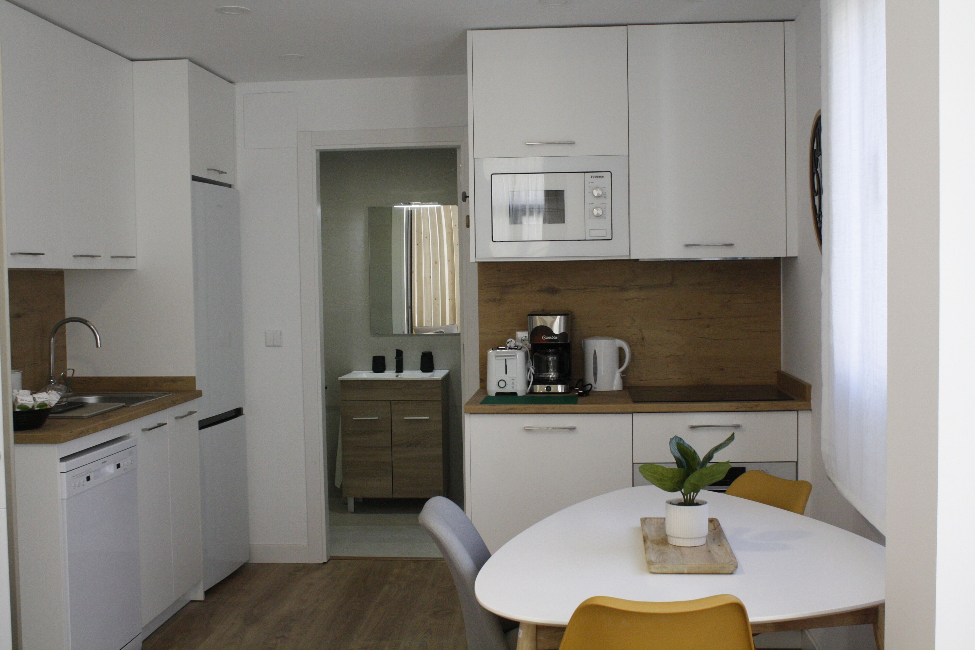 Una cocina compacta con armarios blancos, un microondas, una cafetera y un comedor con una mesa y sillas.