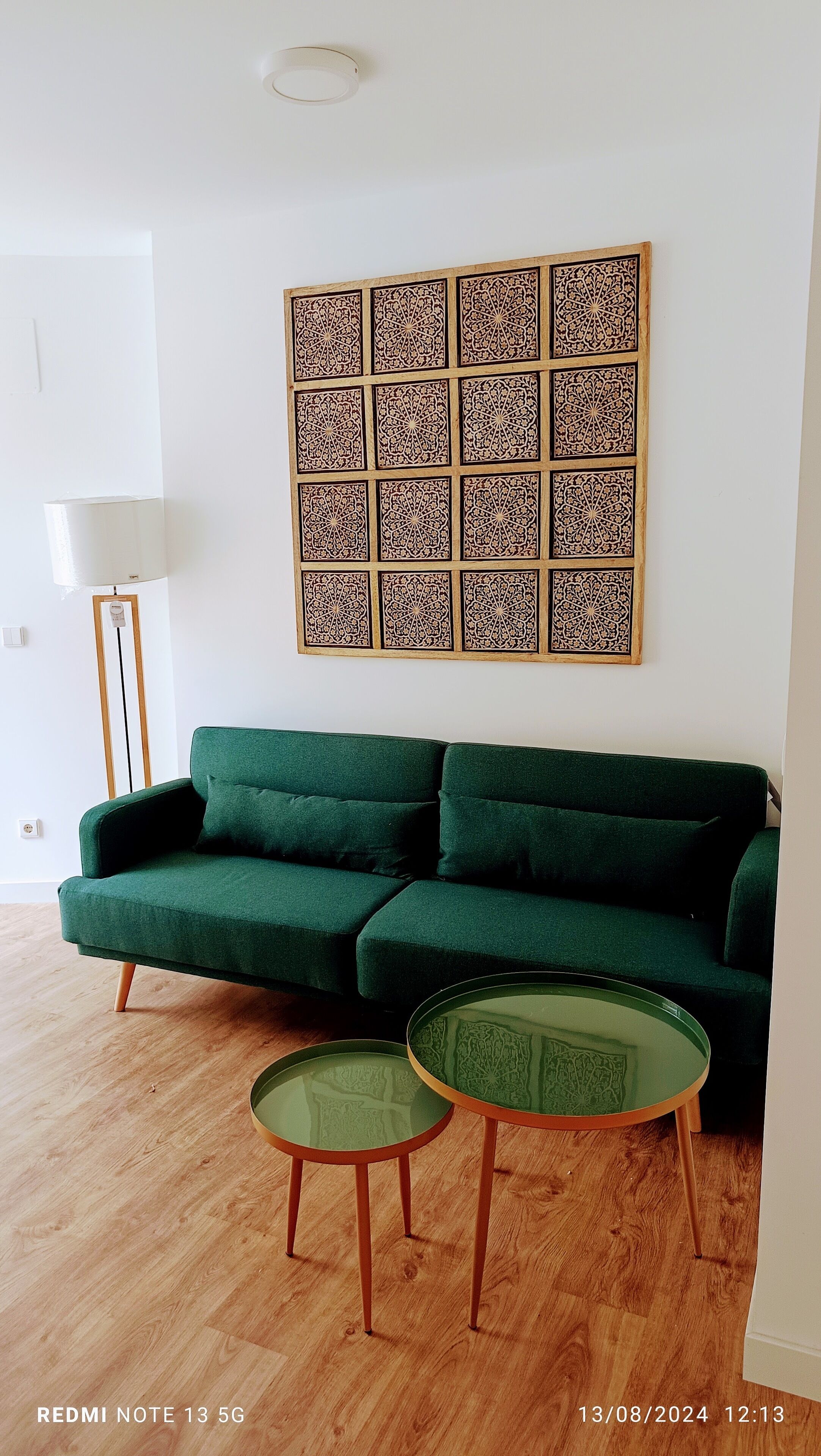 Ett vardagsrum med en grön soffa, två soffbord i glas och en dekorativ panel på väggen.