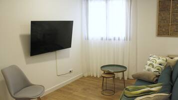 Una sala de estar con un televisor de pantalla plana montado en la pared, un sillón gris, una mesita redonda y un sofá con cojines estampados.