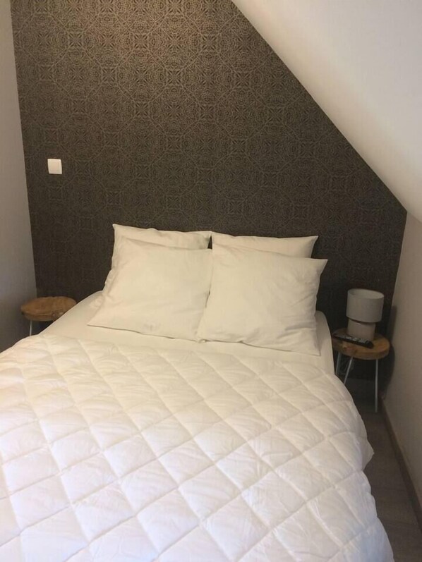 A bed with a quilted white duvet, a patterned wall, a small wooden stool, and a nightstand with a lamp.