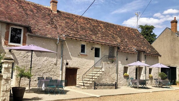 Un bâtiment rustique en pierre, doté d’un toit en tuiles, d’un espace extérieur aménagé avec des parasols et d’un escalier menant à l’entrée.