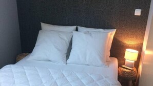 Un lit bien fait, avec une couette blanche matelassée, une tête de lit à motifs foncés et une lampe de chevet avec une bougie allumée.