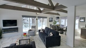 Un vaste salon comprenant un grand canapé, une table basse et un téléviseur fixé au mur. On y trouve également un coin repas avec une table et des chaises, ainsi qu’un coin cuisine avec une chaise rouge.