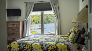 Une chambre avec une grande fenêtre, une commode en bois, un lit recouvert d’une couvre-lit à motifs floraux et une lampe.