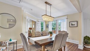 A dining area with a wooden table, upholstered chairs, a chandelier, and a view of the outdoors through large windows. - Historic St Augustine | Courtyard Fireplace | Dog Friendly (St. Augustine Beach)