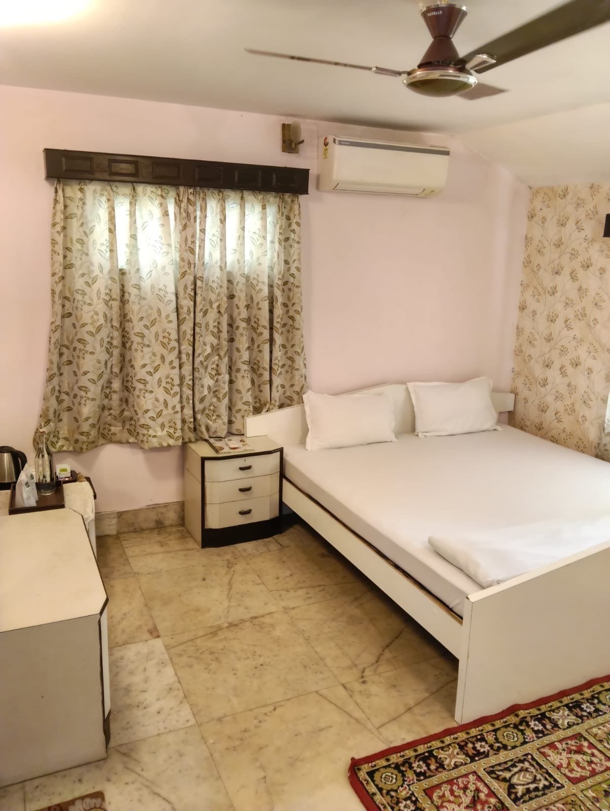 A bedroom with a bed, bedside table, air conditioning unit, curtains, and a ceiling fan.