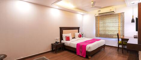 A hotel room with a bed, bedside table, chair, desk, and a window with patterned curtains.