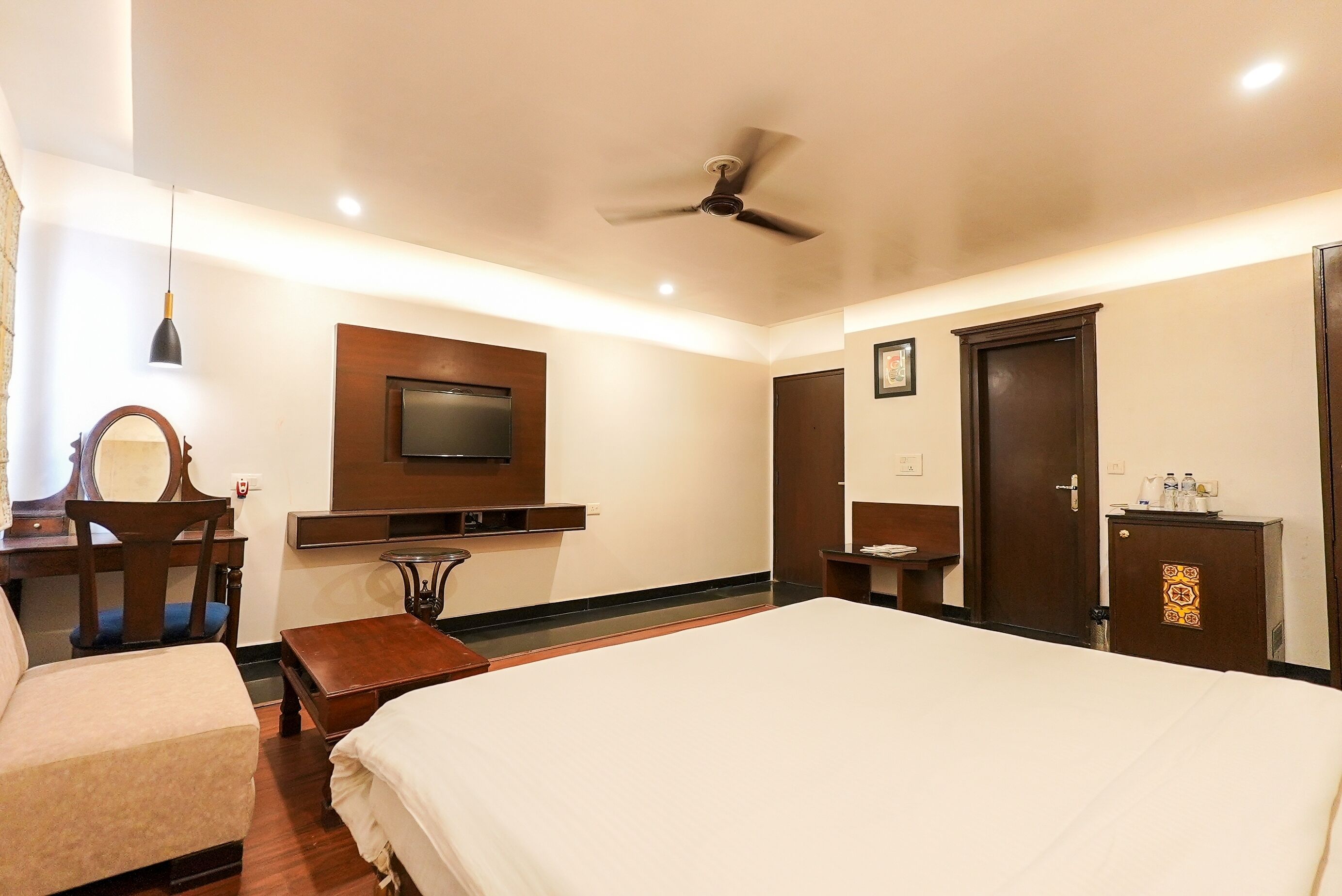 Habitación de hotel con cama, escritorio con silla, televisor montado en la pared, ventilador de techo y armario.