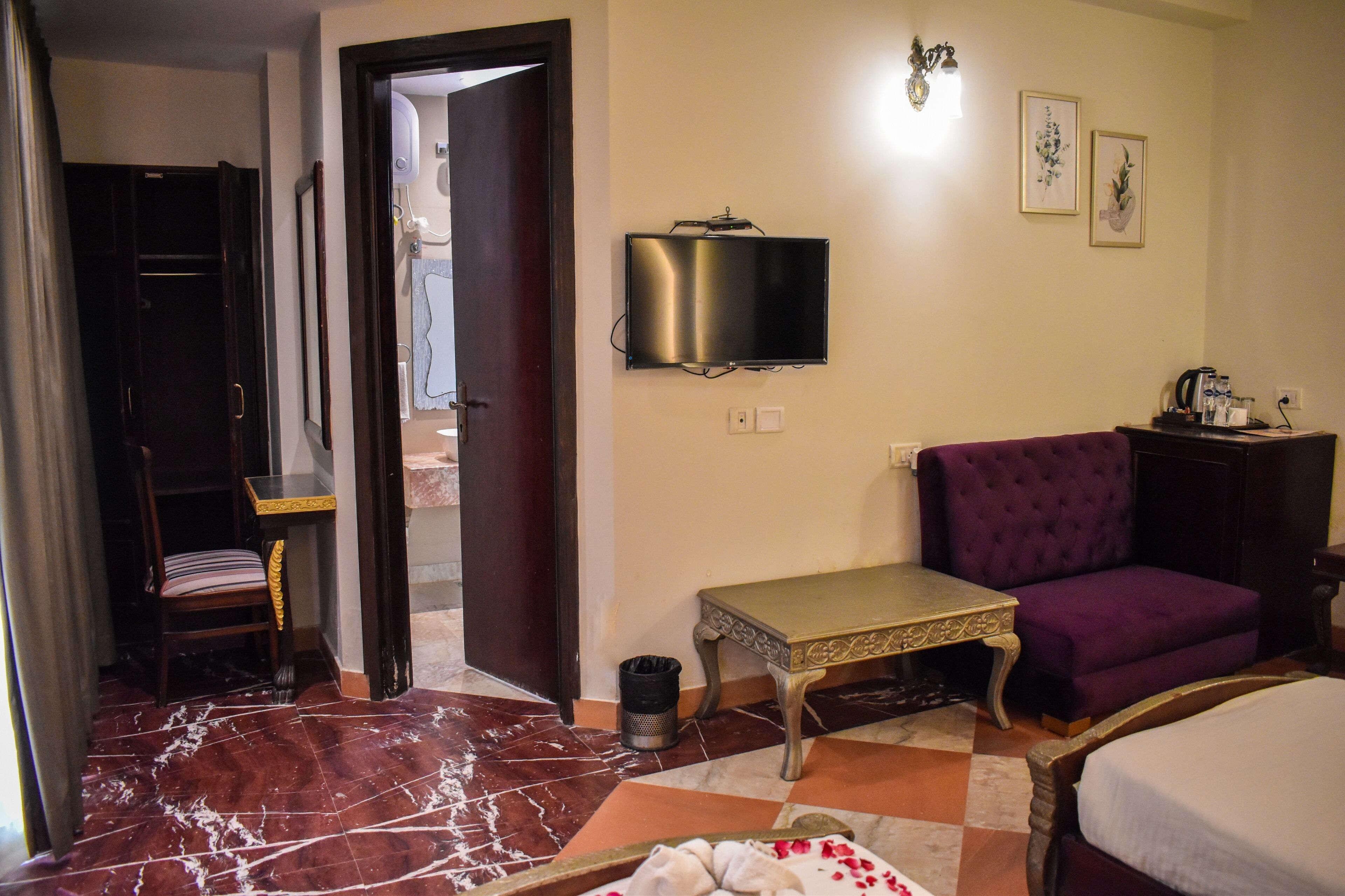 A hotel room with a bed, a purple armchair, a small table, a TV mounted on the wall, and a door leading to another room.