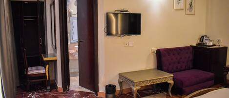A hotel room with a bed, a purple armchair, a small table, a TV mounted on the wall, and a door leading to another room.