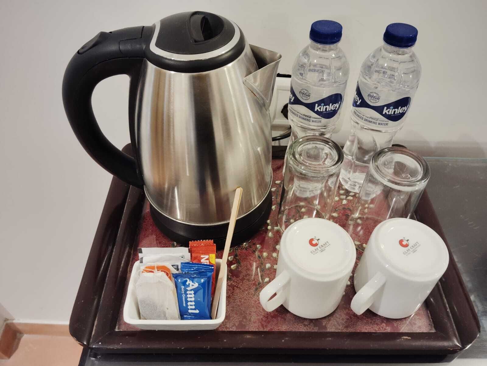 A tray with a stainless steel kettle, two Kinley water bottles, two glass jars, a white mug, a blue packet, and a white mug with a red logo.