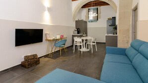 Ein modernes Wohnzimmer mit einem blauen Sofa, einem Fernseher, einem Essbereich mit weißen Stühlen und einer Küche im Hintergrund.