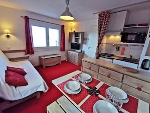 A compact room with a bed, a small table set for dining, a kitchenette, and a window with curtains. - Superior studio cabin near slopes, sleeps 4, south balcony, ski locker (Les Adrets-Prapoutel)