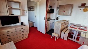 Quarto compacto e bem organizado, com televisão, mobiliário de madeira, cozinha compacta e área de refeições.