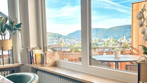 A room with a large window offering a view of a cityscape and mountains. There is a table with chairs, a radiator, and a shelf with books and decorative items.