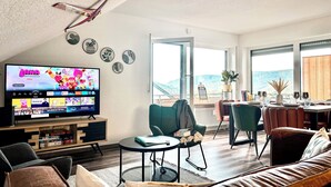 A modern living room with a flat-screen TV, comfortable seating, a dining area, and large windows offering a view of the outdoors.
