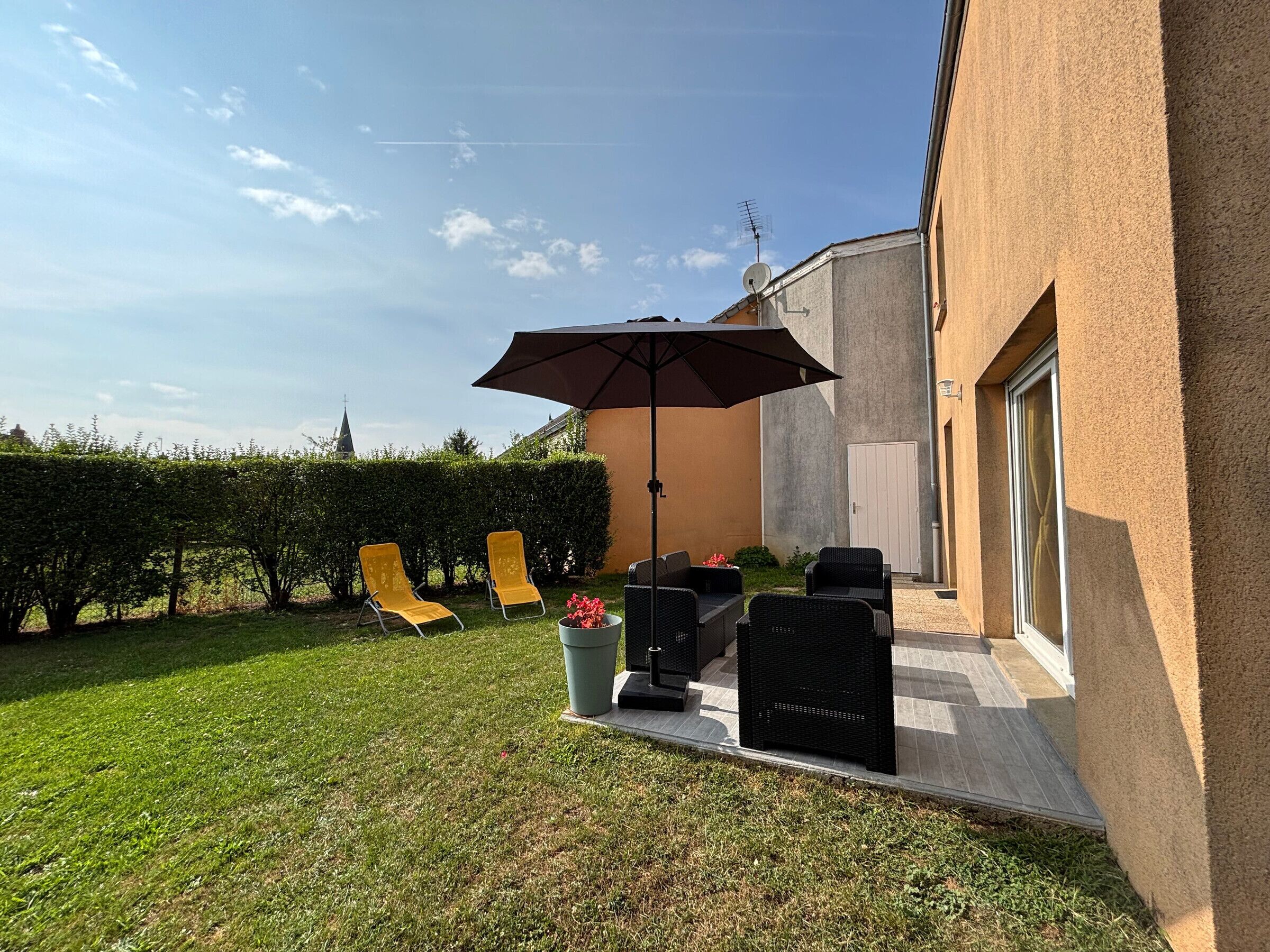 Une terrasse avec des meubles d’extérieur, un grand parasol et une haie.