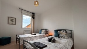 A bedroom with two beds, a window with curtains, a radiator, a bedside table with a lamp, and a hanging light fixture.
