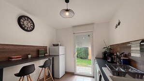 A modern kitchen with a built-in refrigerator, a wooden countertop, two black bar stools, a hanging light fixture, and a door leading to a garden.