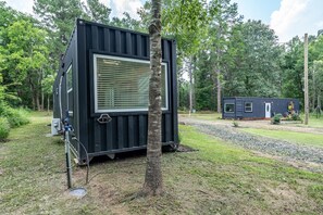 A compact, dark-colored container-style building with a large window, surrounded by trees and a gravel path. - Pod in the Piney Woods (Nacogdoches)
