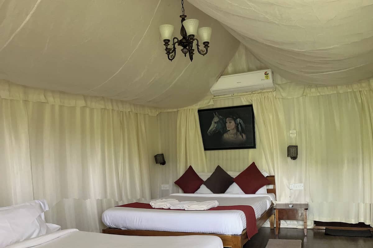 Um quarto amplo coberto por uma tenda, com duas camas de madeira, um lustre e um ar condicionado fixado na parede.