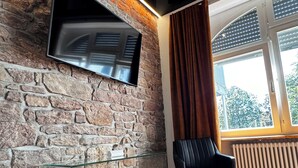 A room with a stone wall, a mounted TV, a glass table, a leather chair, and a large window with curtains.
