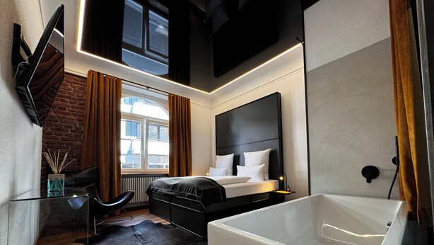 A modern bedroom with a large bed, a separate bathroom with a bathtub, a window with curtains, and a brick wall.