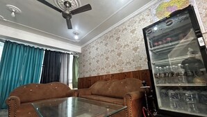 A living room with a sofa, glass coffee table, ceiling fan, patterned wallpaper, and a refrigerator with beverages. - Hotel Honey N Spice (Dharamshala)