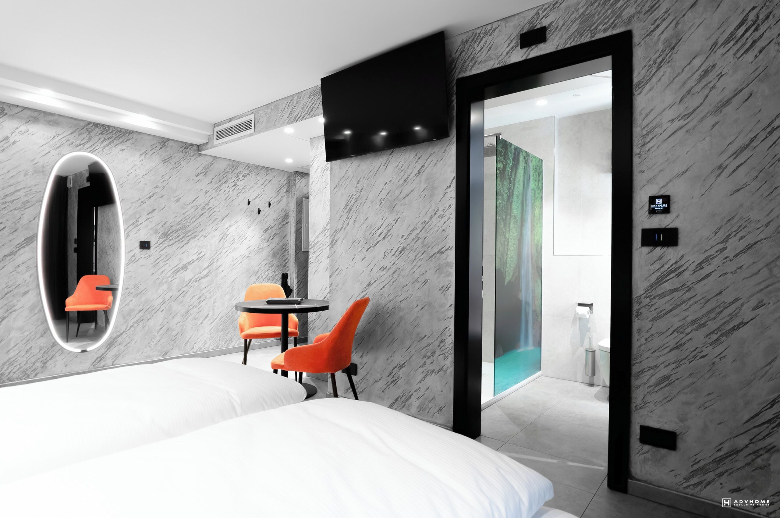 A modern hotel room with a large bed, a seating area with orange chairs, a small table, a TV mounted on the wall, and a bathroom visible through an open door.