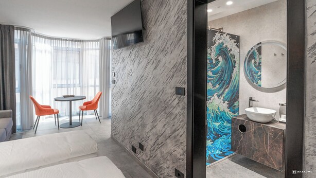Une chambre d’hôtel moderne avec un mur en marbre, une table ronde avec deux chaises, une grande fenêtre avec des rideaux, et une salle de bain avec un mur au motif ondulé original et un miroir rond.