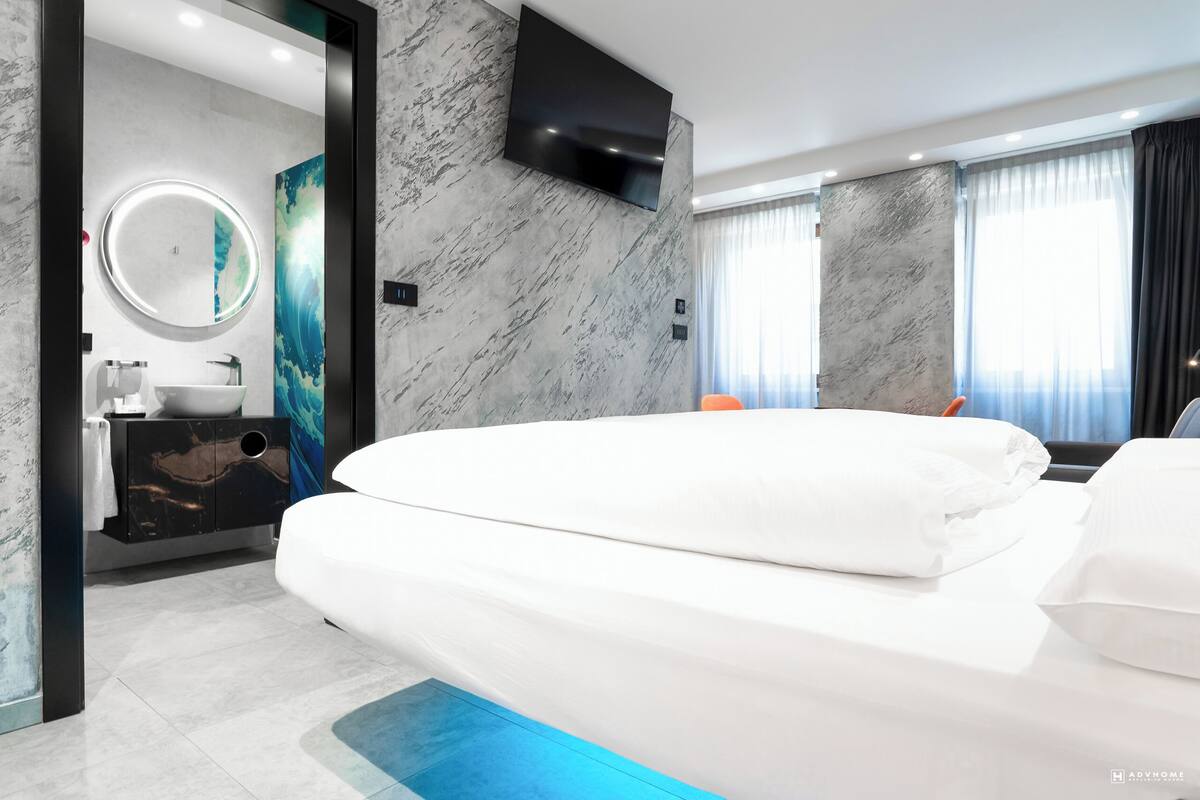 A modern hotel room with a large bed, a round mirror, a TV mounted on the wall, and a bathroom with a marble sink and vanity.