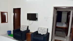 A hotel room with two dark blue armchairs, a small wooden table, a TV mounted on the wall, a bed, and a bathroom visible through an open door. - Krishnam Guest House (Gwalior)