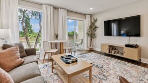 A modern living room with a sofa, coffee table, TV, and a view of greenery through large windows. - Renovated Chic, Riverfront Condo w/ Pool (Nashville)