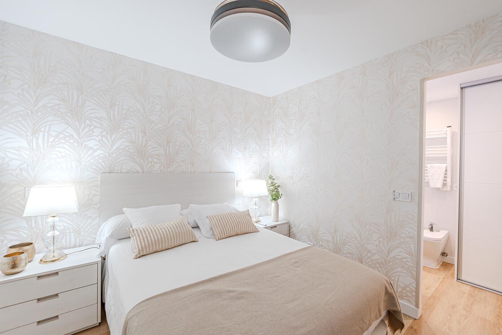 A bedroom with a bed, bedside tables, a nightstand, a lamp, and a bathroom visible through an open door.
