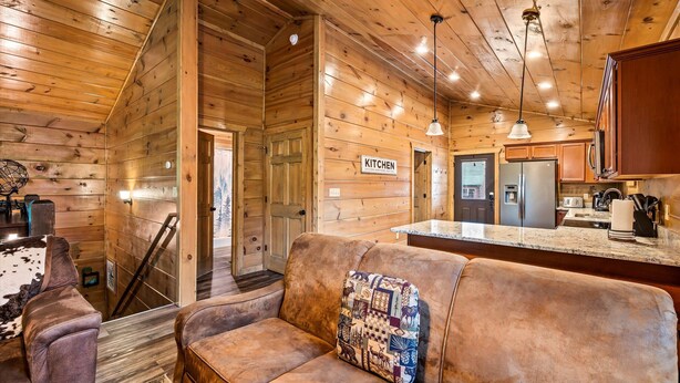Interior acogedor de una cabaña con detalles de madera, un sofá con un almohadón acolchado, una zona de cocina con refrigerador y fregadero, y un letrero que indica la cocina.
