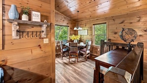 Interior de una cabaña de madera con una mesa de comedor, sillas, una estantería con objetos decorativos y un letrero que dice 'STAY humble'.