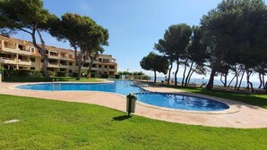 A swimming pool with a surrounding grassy area and trees, adjacent to a multi-story building. - Alcanar Costa (CT)