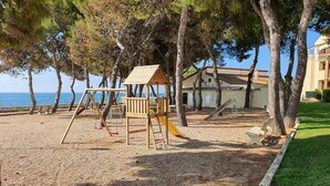 A playground with a swing set and a slide, surrounded by trees and a building in the background. - Alcanar Costa (CT)