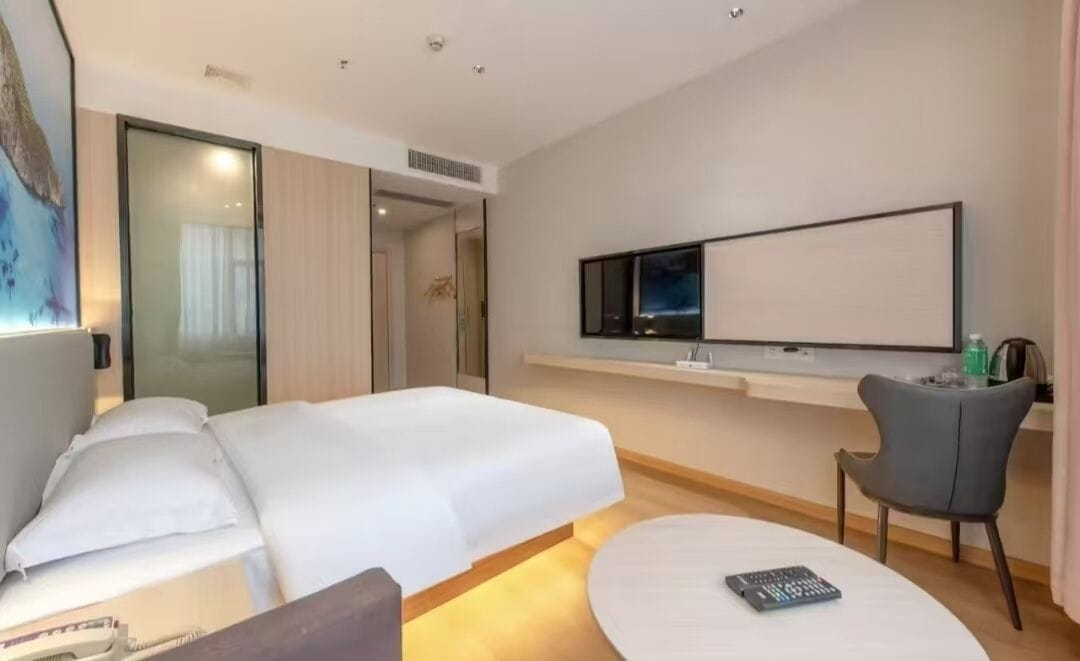 Ein modernes Hotelzimmer mit einem großen Bett, einem flachbildfernseher an der Wand, einem kleinen Schreibtisch mit Stuhl und einem Nachttisch mit Fernbedienung.