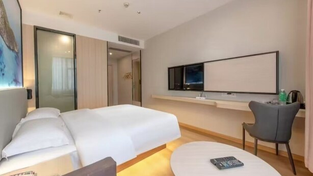 A modern hotel room with a large bed, a flat-screen TV mounted on the wall, a small desk with a chair, and a bedside table with a remote control. - Manli Hotel (Foshan)