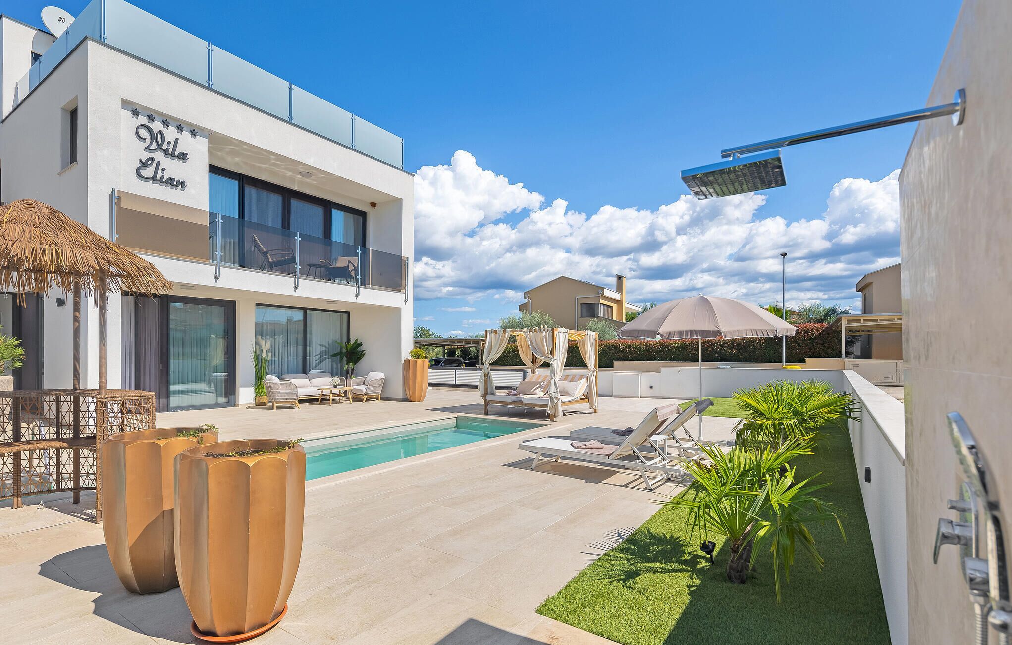 Area piscina moderna all'aperto con poltrone relax, un gazebo e un ombrellone grande.