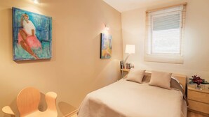 Quarto com cama, mesa de cabeceira, luminária e duas pinturas na parede.