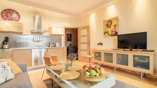 Uma sala de estar moderna com uma mesa de centro de vidro, um sofá, uma televisão e uma cozinha ao fundo.