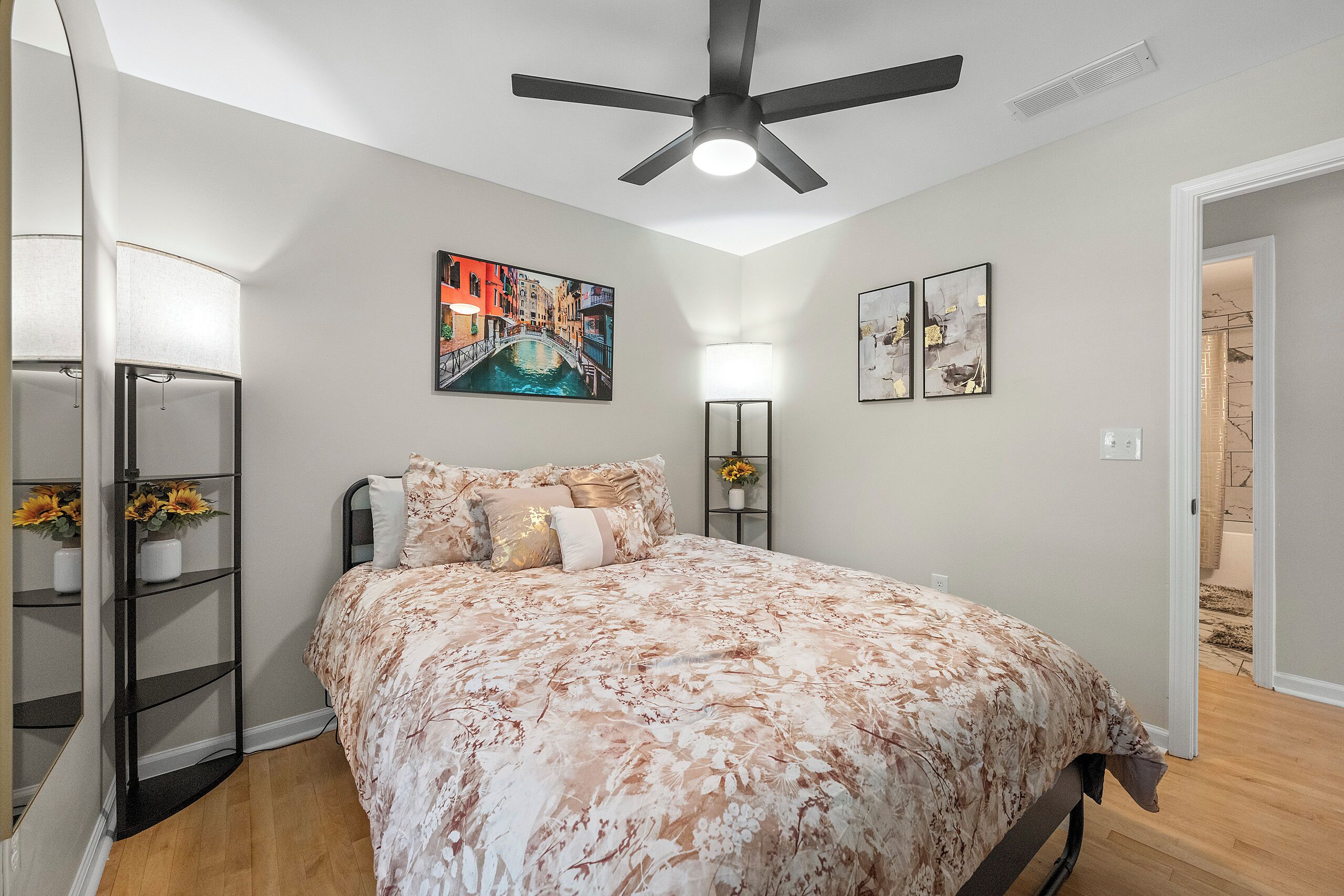 A bedroom with a bed, a ceiling fan, a painting of a canal, and a bookshelf.